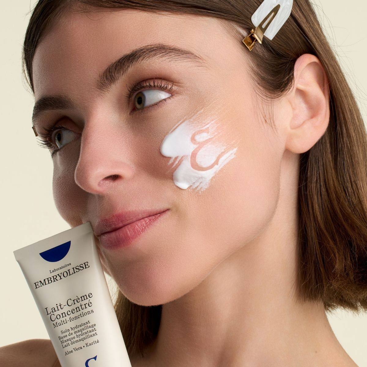 Model with a swatch of Embryolisse Lait-Creme Concentre on her cheek, holding the tube; official maker photograph