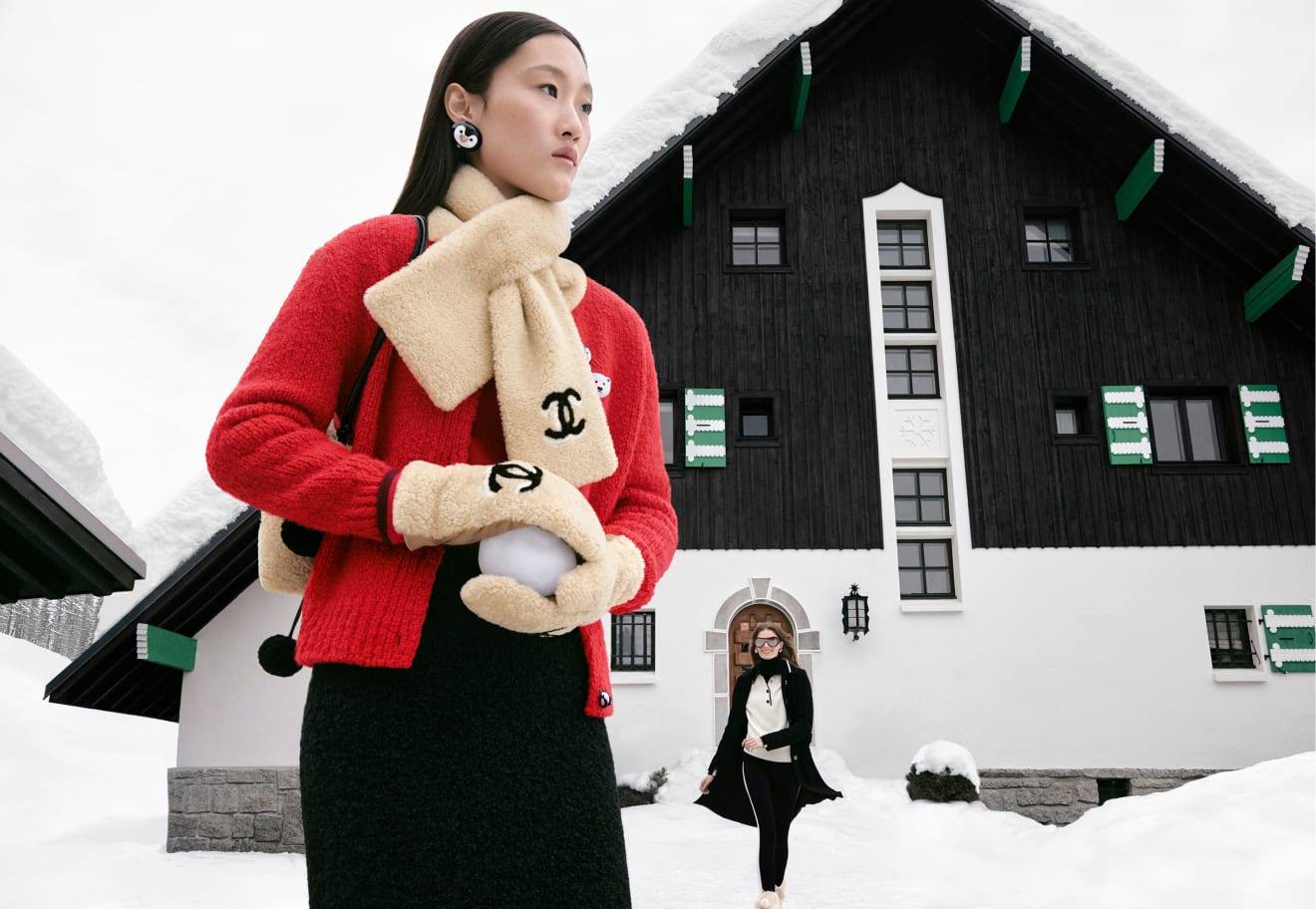 A model in a red Chanel Coco Neige 2026 cardigan with cream shearling scarf and mittens, holding a snowball outside a chalet.