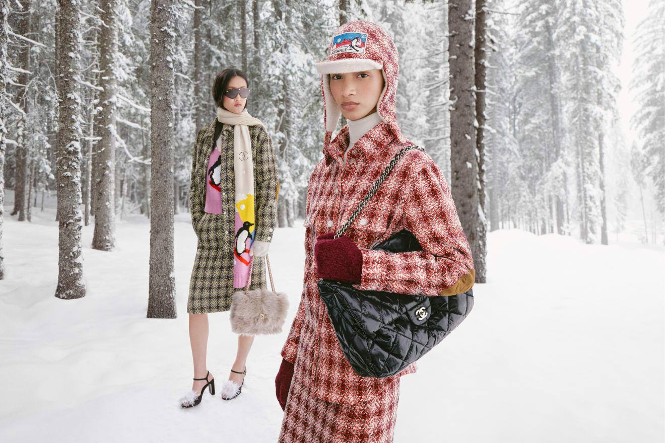 Two models in checked Chanel Coco Neige 2026 tweeds in a snowy forest, one carrying a black quilted maxi flap bag.